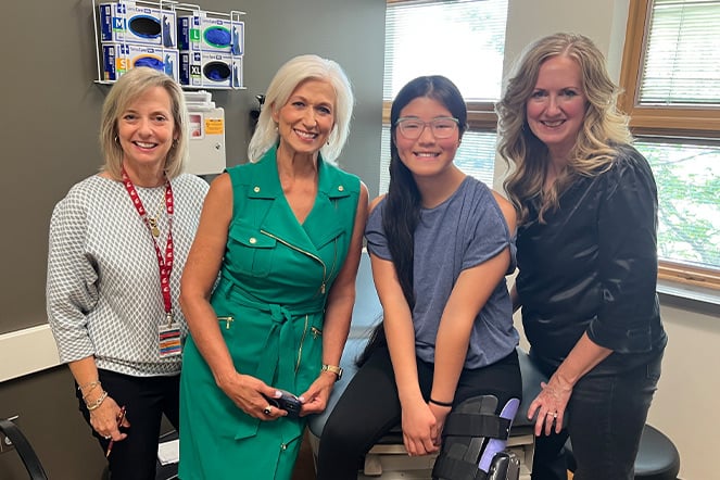 Four people pose together in a clinical exam room, with a patient seated on an examination table wearing glasses and a leg brace.