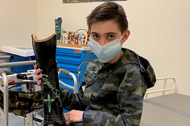 Young patient wearing a face mask and camouflage hoodie sits in a clinical therapy room while holding a black prosthetic limb upright. Medical equipment, therapy supplies, and a colorful bead maze activity toy are visible in the background. Writing and a green ribbon graphic appear on the surface of the prosthetic limb.