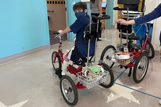 Young patient wearing a mask rides a red adaptive tricycle through a hospital therapy area. The tricycle features supportive seating and a backrest, with a small character toy in the front basket. A clinician walks alongside with a hand extended toward the tricycle, while a second adaptive tricycle is visible nearby in the brightly lit hallway.
