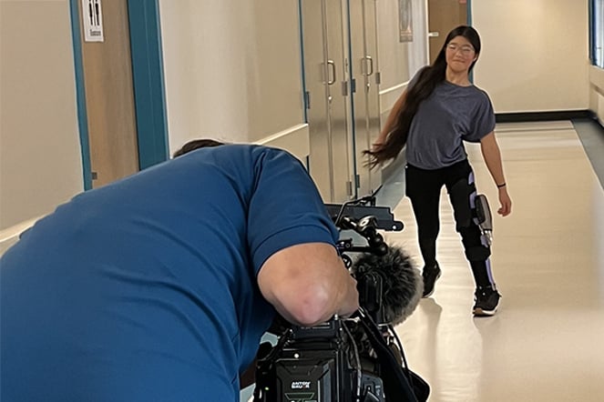A patient wearing a specialized leg brace walks down a hospital hallway while being filmed by a videographer operating a professional video camera.