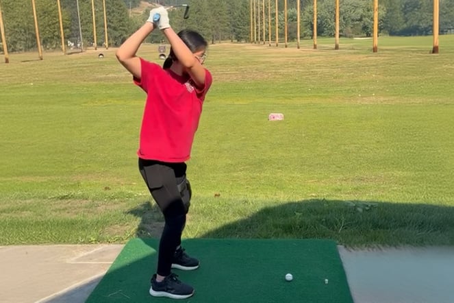 A patient practices a golf swing at a driving range while standing on a hitting mat and holding a golf club overhead during the backswing.