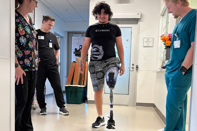 A patient wearing a lower-limb prosthesis walks down a hospital hallway while several clinicians observe. The patient wears a black T-shirt and patterned shorts, and the prosthetic leg is clearly visible during a mobility assessment. Healthcare professionals in scrubs stand on either side of the corridor, with exam rooms and clinical equipment visible in the background, highlighting rehabilitation and prosthetic care.