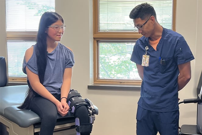 A patient sits on an examination table wearing a specialized leg brace while speaking with a healthcare provider in a clinical exam room.