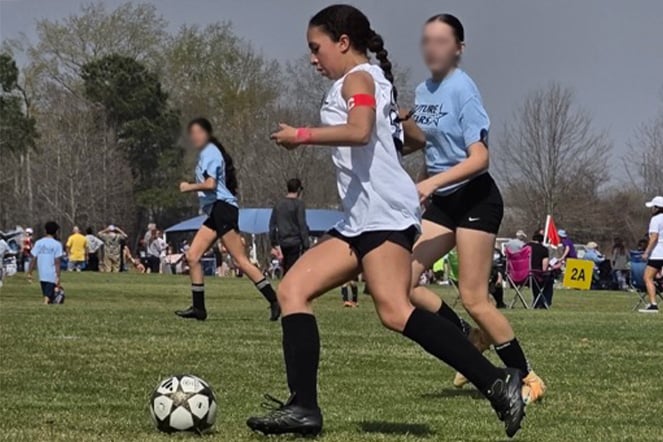 A youth soccer player wearing a white jersey, black shorts and black knee-high socks dribbles a soccer ball across a grassy field during an outdoor match. Other players in light blue jerseys run nearby as play continues around the field.