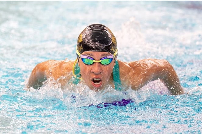 A competitive swimmer performs the butterfly stroke during a race in an indoor pool. The swimmer wears a swim cap and mirrored goggles while emerging from the water with both arms extended outward. Water splashes around the swimmer as they move through the lane, highlighting the intensity and athleticism of the competition.