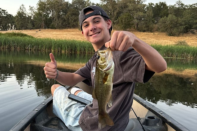 Teenager sitting in a small boat holds up a fish while giving a thumbs-up gesture. The boat floats on calm water bordered by tall reeds and grassy shoreline, with trees in the background.