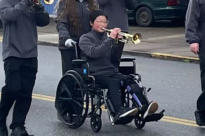 A patient seated in a wheelchair plays a trumpet while participating in a community parade or outdoor marching event.