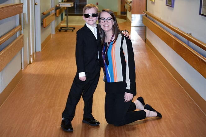 A child wearing a suit and sunglasses smiles with an adult woman kneeling next to him.