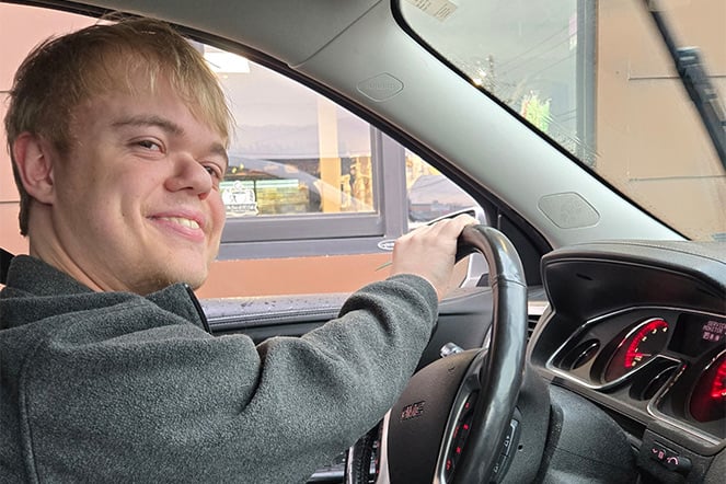 a smiling teenager in the driver's seat of a car waiting for food at a drive thru window
