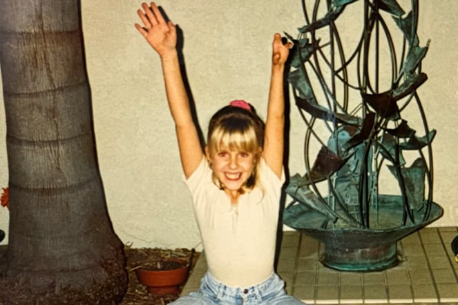 A young girl sits on the ground in a split position with her arms raised triumphantly above her head
