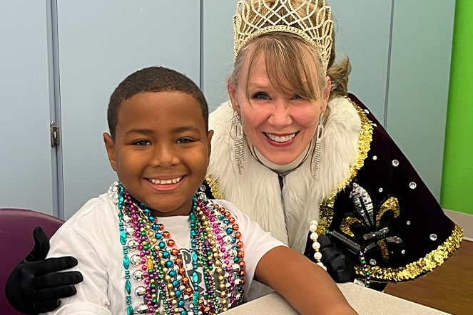 a child with multiple plastic bead necklaces smiling next to a woman in a tiara and royal cape