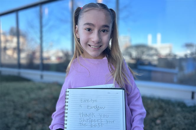  a young girl holding a notebook that reads "Evelyn. Thank you Shriners, you're the best." in her own handwriting