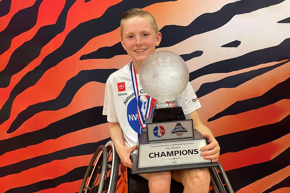 International Patient Ambassador Brandon in a wheelchair holding a wheelchair basketball championship trophy