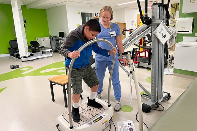 a child stands on a balancing device during a physical therapy session while a physician in hospital scrubs watches on