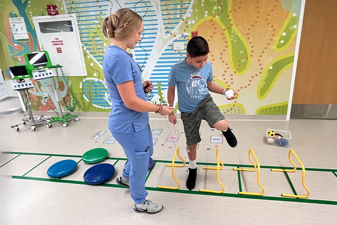 a child side-stepping over obstacles in a physical therapy while a physician in hospital scrubs watches on