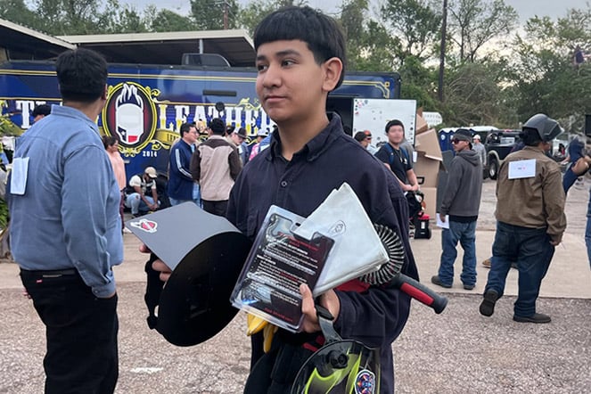  a teenager holding welding supplies outside in a dirt parking lot surrounded by people