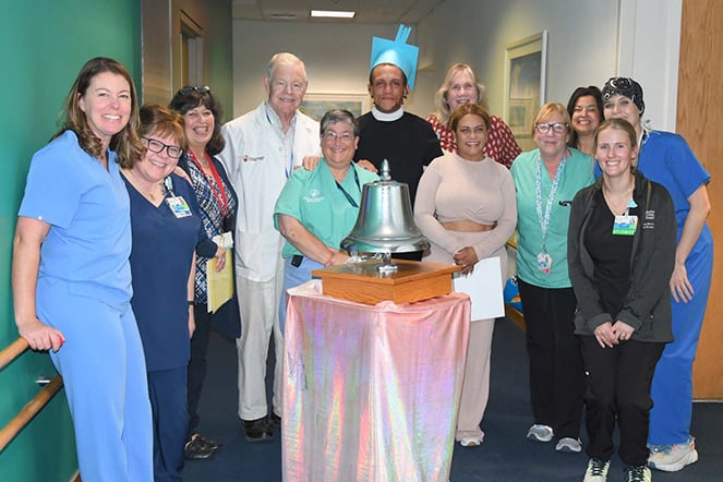 burn patient with a paper graduation cap surrounded by Shriners Children's Boston staff in front of a metallic bell on a table