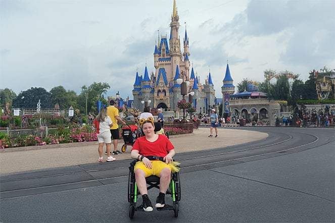 Cerebral palsy patient sitting and smiling in their wheelchair in front of Cinderella's castle at Disney World