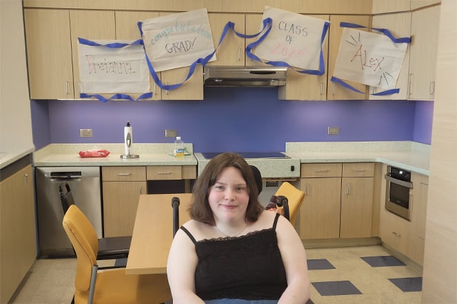 Cerebral patient sitting and smiling in front of signs that read "Breianna" "Congratulations, Grad" "Class of 2024" and "Alex"