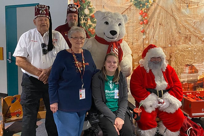 Cerebral palsy patient smiling for a photo with with two Shriners in fezzes, a woman, a polar bear mascot and Santa Claus 