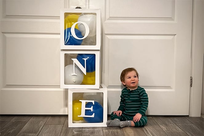 an infant sitting on the ground next to clear boxes containing balloons stacked in order to spell the word "ONE"