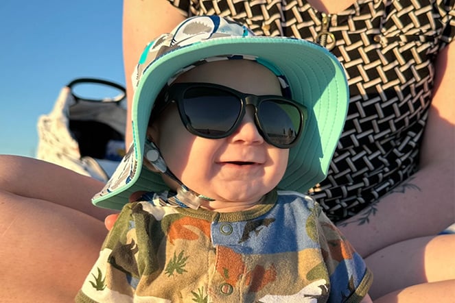 a smiling infant wearing sunglasses and a bucket hat at a beach