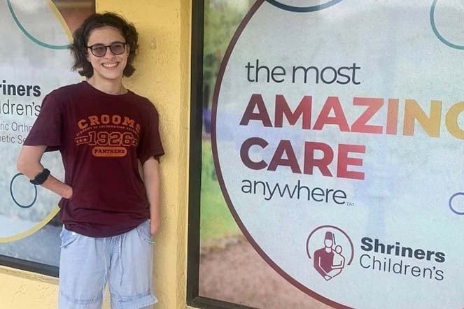 Grace standing in front of Pediatric Orthotic and Prosthetic Services facility, The Most Amazing Care Anywhere logo, Shriners Children's logo
