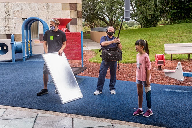 a young girl with a leg prosthetic on a photoshoot set with two men holding camera and lighting gear