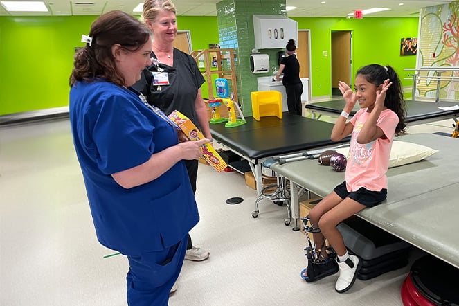 a physician wearing hospital scrubs smiling and looking at a small child as she makes a silly face back at her