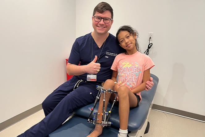 A physician in hospital scrubs giving a thumbs up next to a smiling child wearing an external fixator on her leg.