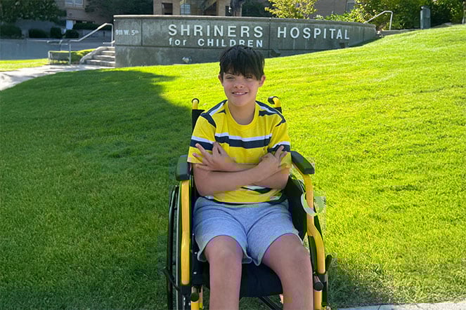 a child in a wheelchair in front of a sign that reads "Shriners Hospital for Children"