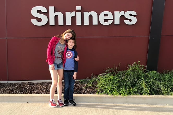 female patient with her arm around her younger brother in front of a sign that reads "Shriners"