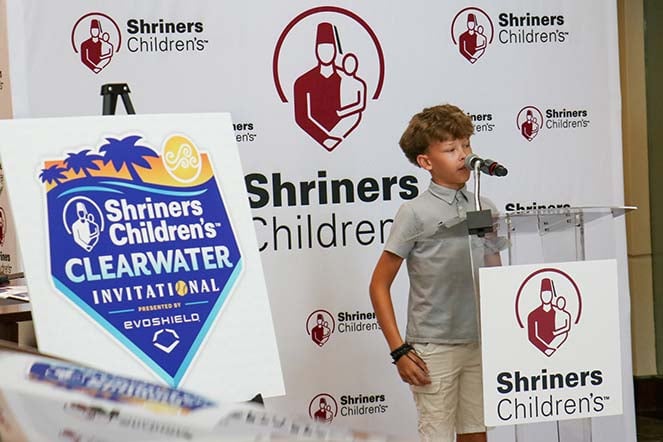 orthopedic patient Zavi speaking at podium, Shriners Children's logo, Shriners Children's Clearwater Invitational presented by EvoShield logo