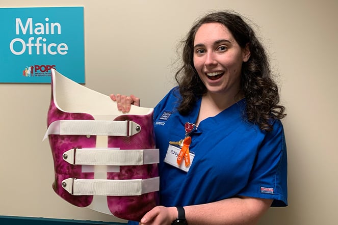 a woman in hospital scrubs holding up a back brace in front of a sign that reads "Main Office"