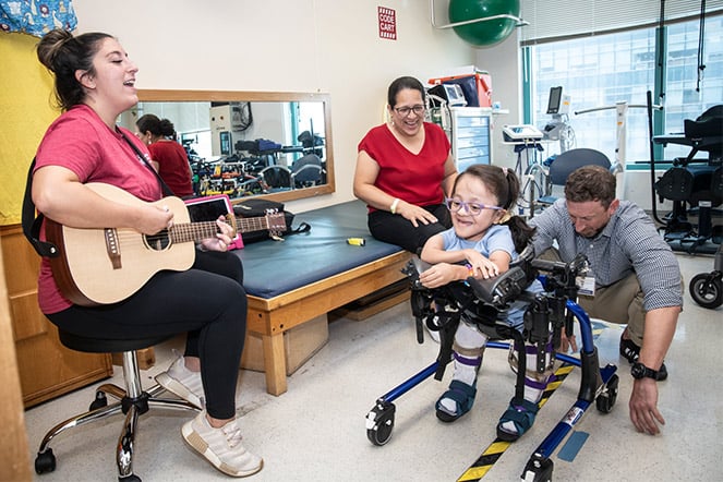an adult woman playing guitar for a patient in a walker and two other adults