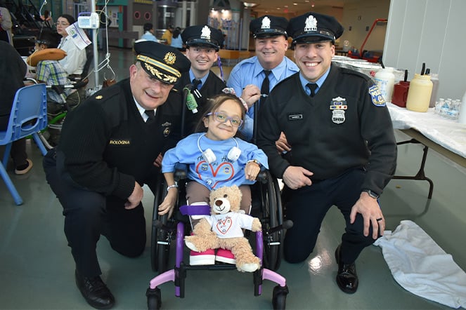 a child in a wheelchair holding a teddy bear surrounded by four smiling uniformed police officers