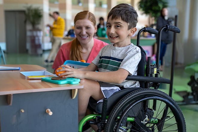 a child in a wheelchair smiling at a table as an adult is crouched near them