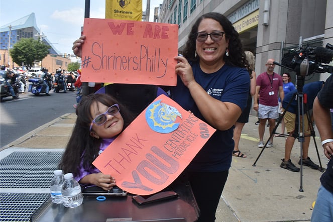 a child in a wheelchair and an adult woman outside holding two signs that read "We Are #ShrinersPhilly" and "Thank You Centurions Motorcycle Club"