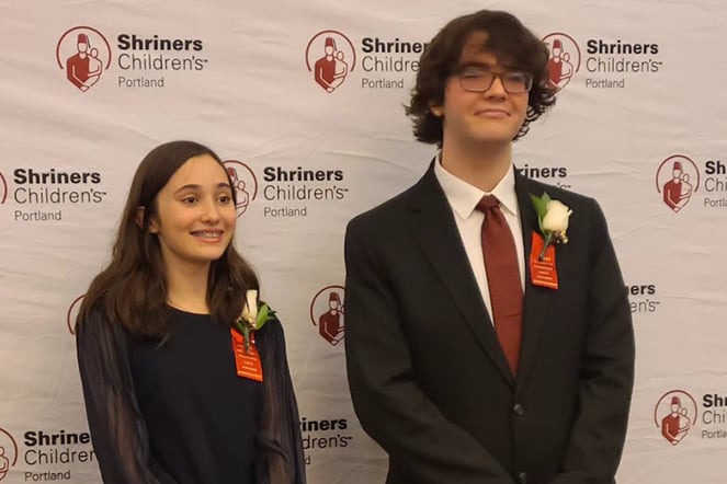 two teenagers in business attire in front of a step and repeat photo background featuring the Shriners Children's Portland logo