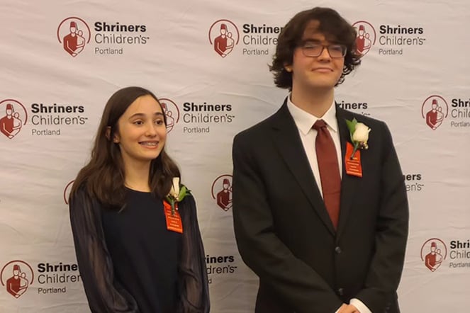 two Patient Ambassadors smiling in front of a photo backdrop with a repeating pattern that features the Shriners Children's logo and reads "Shriners Children's Portland"