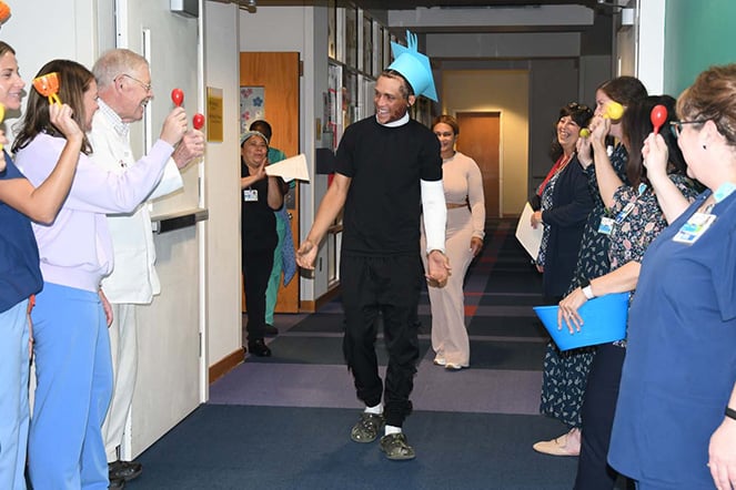 Patient Leufry wearing a paper graduation cap walking down a hallway lined with staff clapping, cheering and ringing bells