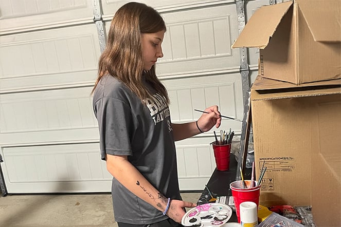 Patient Angel painting a canvas against cardboard boxes in a garage