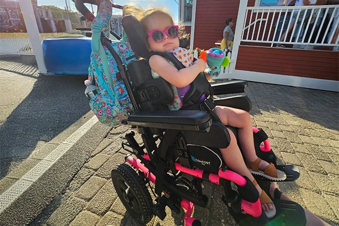 a young child wearing sunglasses sitting in a powerchair against a sunny sky