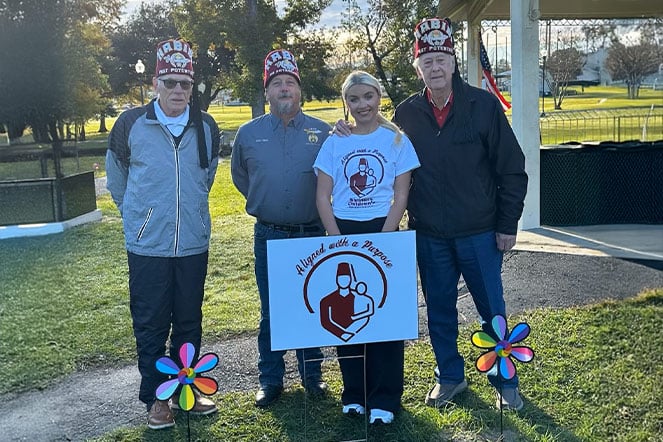 Patient Anna with three members of the Habibi Shriners outside in front of a sign with the Shriners Children's logo that reads "aligned with a purpose"