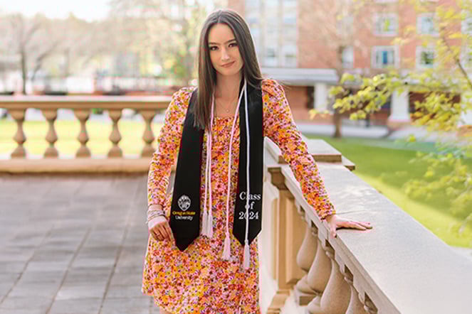 Peighten in a floral dress on a balcony wearing honors cords and a graduation sash that reads "Oregon State University Class of 2024" 