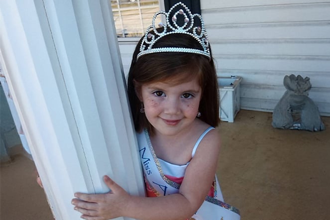 a young girl wearing a sash and tiara standing halfway behind a column