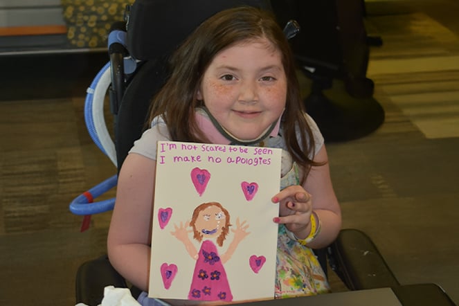 a child sitting in a wheelchair holding up a painting of a young girl with hearts around her under the words "I'm not scare to be seen I make no apologies"