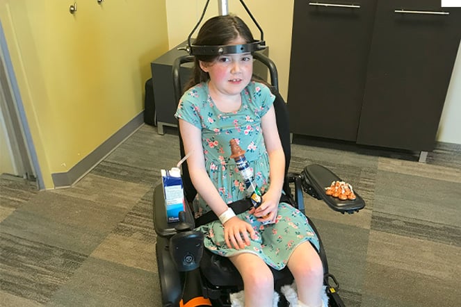 a young girl with a halo traction device sitting in a wheelchair with a tube of chocolates