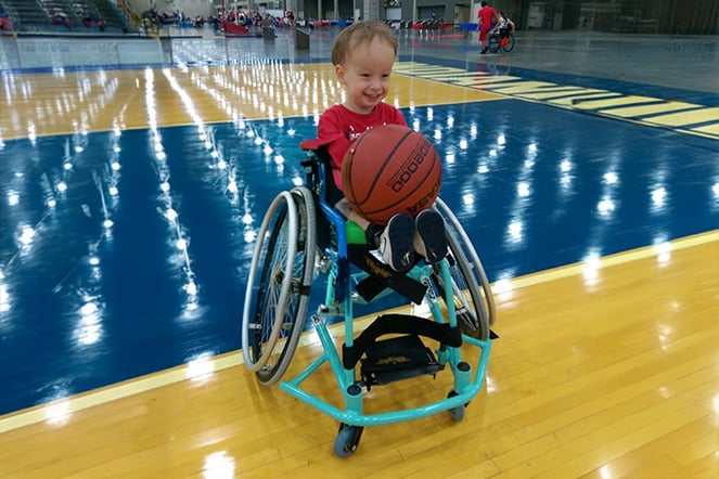 A young spina bifida patient in a wheelchair holding a basketball on a basketball court