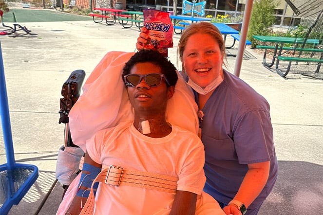 a child in a power chair wearing sunglasses sits with two adults under the shade of an umbrella